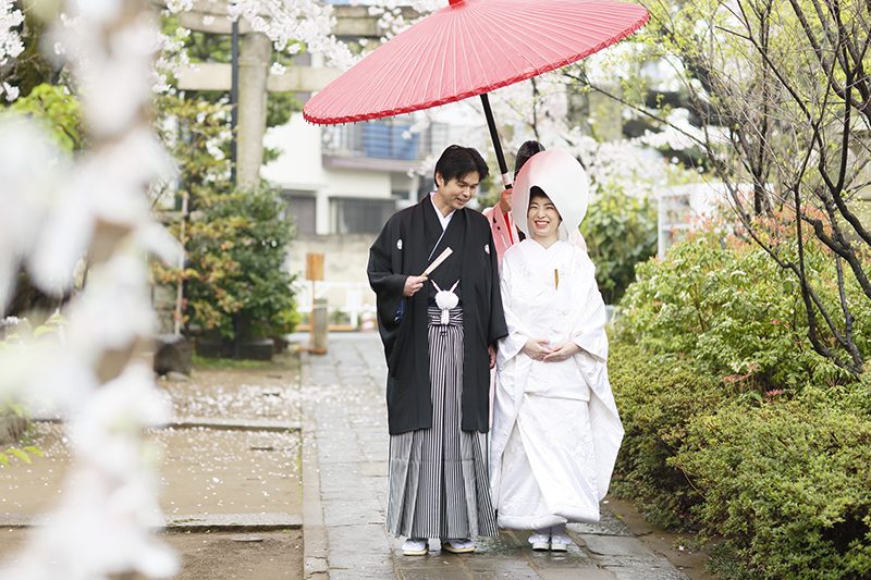 「桜の季節の結婚式。当日はあいにくの雨でしたが、傘をさして、合間合間で手早く撮影しました。カメラマンや着付け・メイクなど本当に慣れたチームだからこそできること。白無垢おかつらの日本の花嫁姿が緑にしっとりと映えて、とても美しい写真になっています。大人世代のふたりだからこそ、紋服と白無垢に日本髪という伝統的なスタイルがよくお似合い。挙式後、新婦はご自分の訪問着に着替えて、ホテルでの会食へ向かわれました。アントワープブライダルでは、こうした挙式後のお引上げまで責任もってお手伝いしています」。
