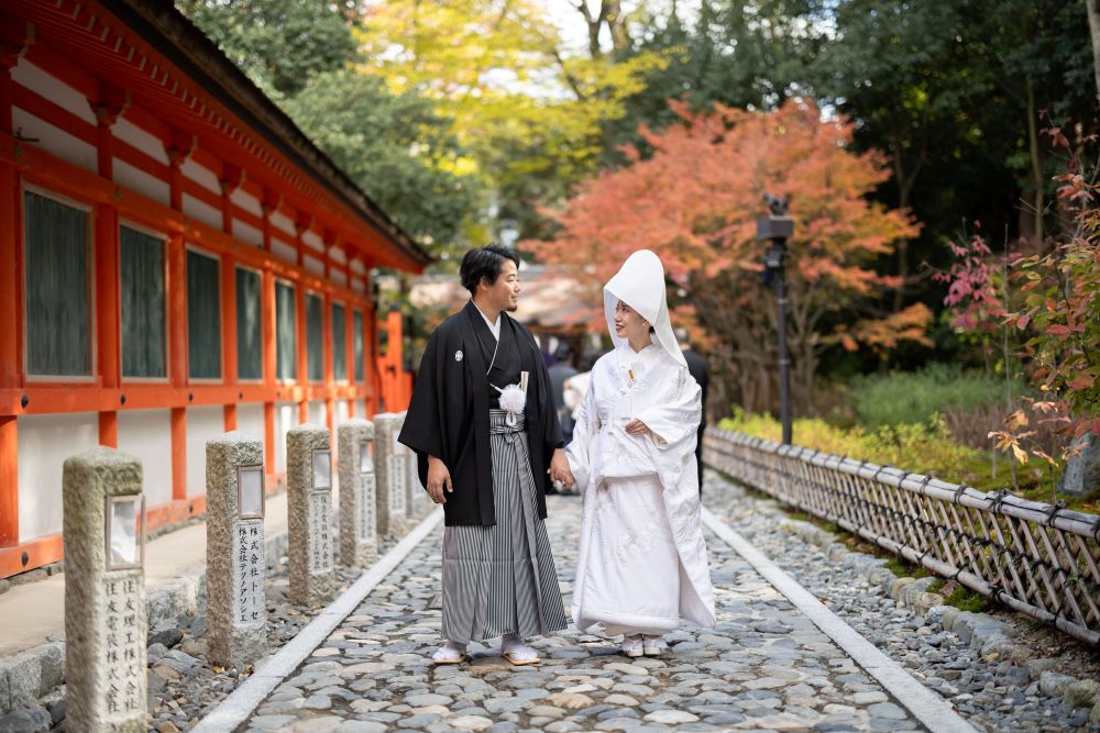 京都らしい挙式がしたいと式場探しをスタート。新婦の出身大学が近くにあり、新郎や家族、ゲストとの思い出が詰まった下鴨神社を選んだ。白無垢に袖を通し、色づく紅葉に包まれる参進、重要文化財の神殿での神聖な儀式に、温かな家庭を築いていく実感と覚悟が生まれた。挙式は親族中心のため、披露宴から参列するゲストにも和を感じる演出を用意。「結婚証明書」と新婦母が書いた御朱印帳に、受付でメッセージを書いてもらった。八坂の塔を望む披露宴会場では、旬を味わう絶品料理でおもてなし。音楽が心に響く祝宴にしたいと曲も厳選。ドラマの主題歌やゲストとの思い出ソングで懐かしい瞬間にタイムスリップし、輝かしい未来へ向けた明るい曲で幕を閉じた。結婚記念日には、挙式当日のような美しい紅葉を眺め、世界遺産の神社と京都の味を堪能できる思い出の場所へ帰ってくることを楽しみにしている。
